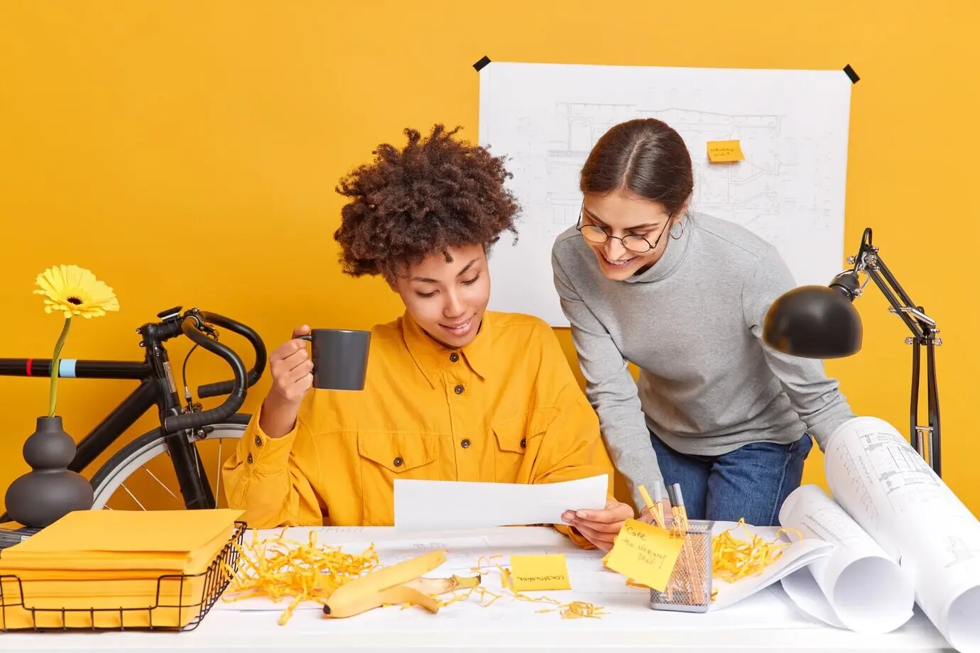 Alegres compañeras de trabajo de raza mixta discuten ideas para el proyecto, comparten opiniones y se preparan para la presentación de bocetos arquitectónicos de un nuevo edificio, posando en el escritorio con papeles alrededor.